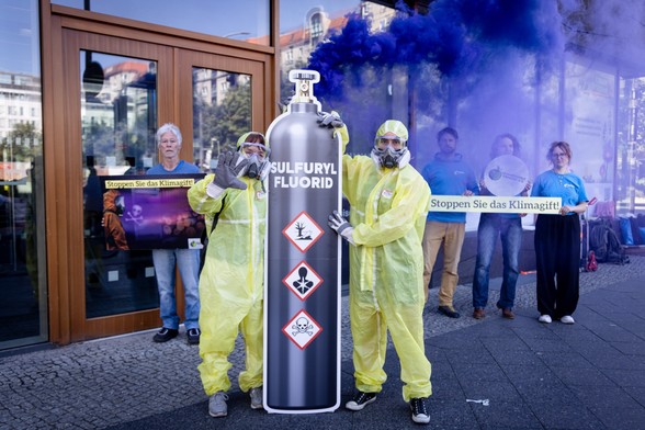 Personen mit Schutzanzügen und -Masken und einer riesigen Abbildung einer Sulfuryl-Fluorid-Flasche, + Schild 